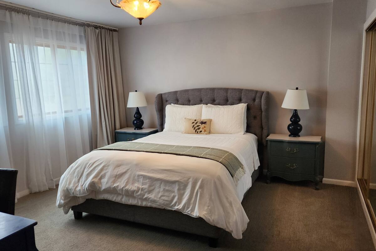 A neatly made bed with a grey headboard, two bedside tables with lamps, a chair, and a window with sheer curtains.