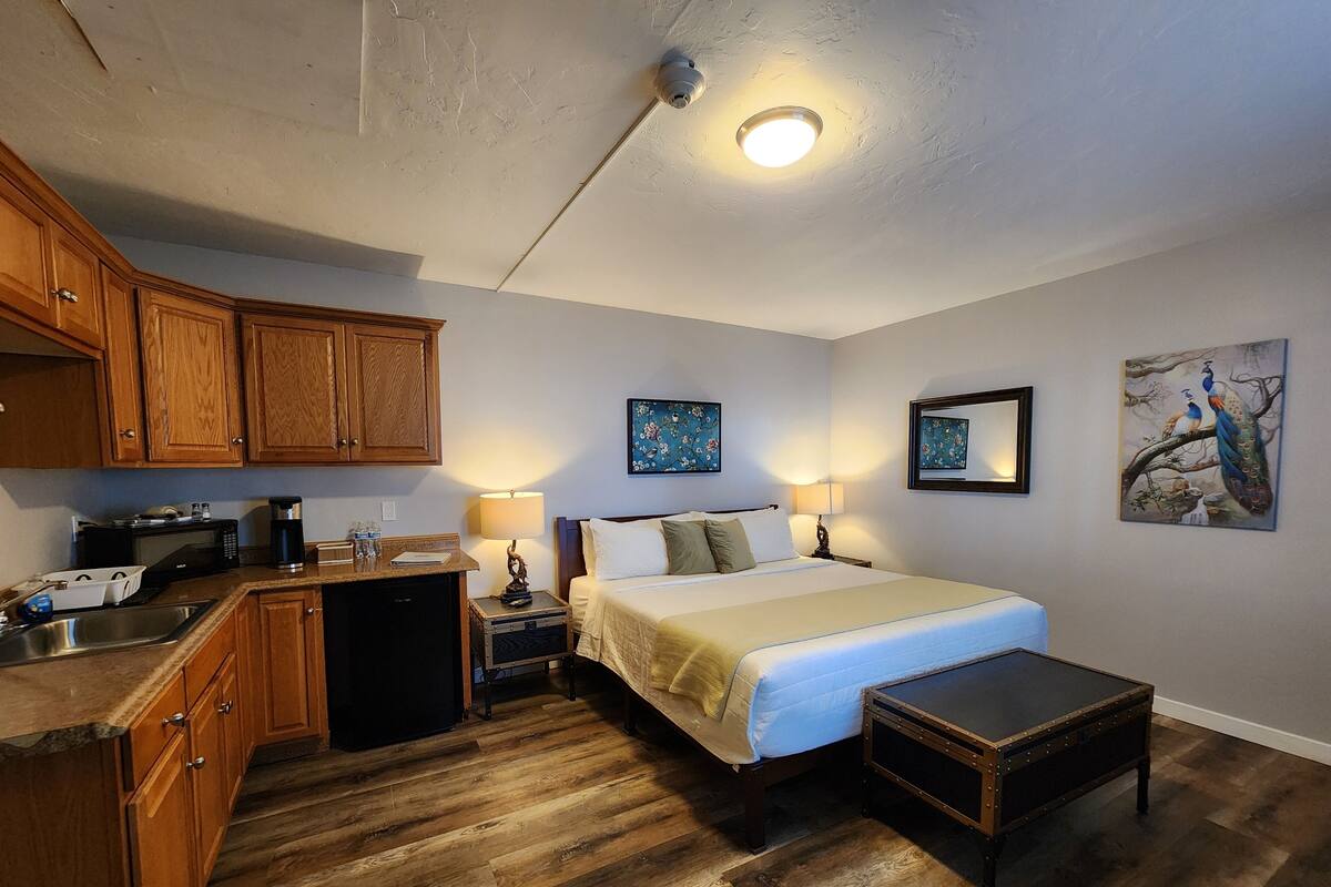 A compact hotel room with a kitchenette, a bed, bedside lamps, and artwork on the walls.