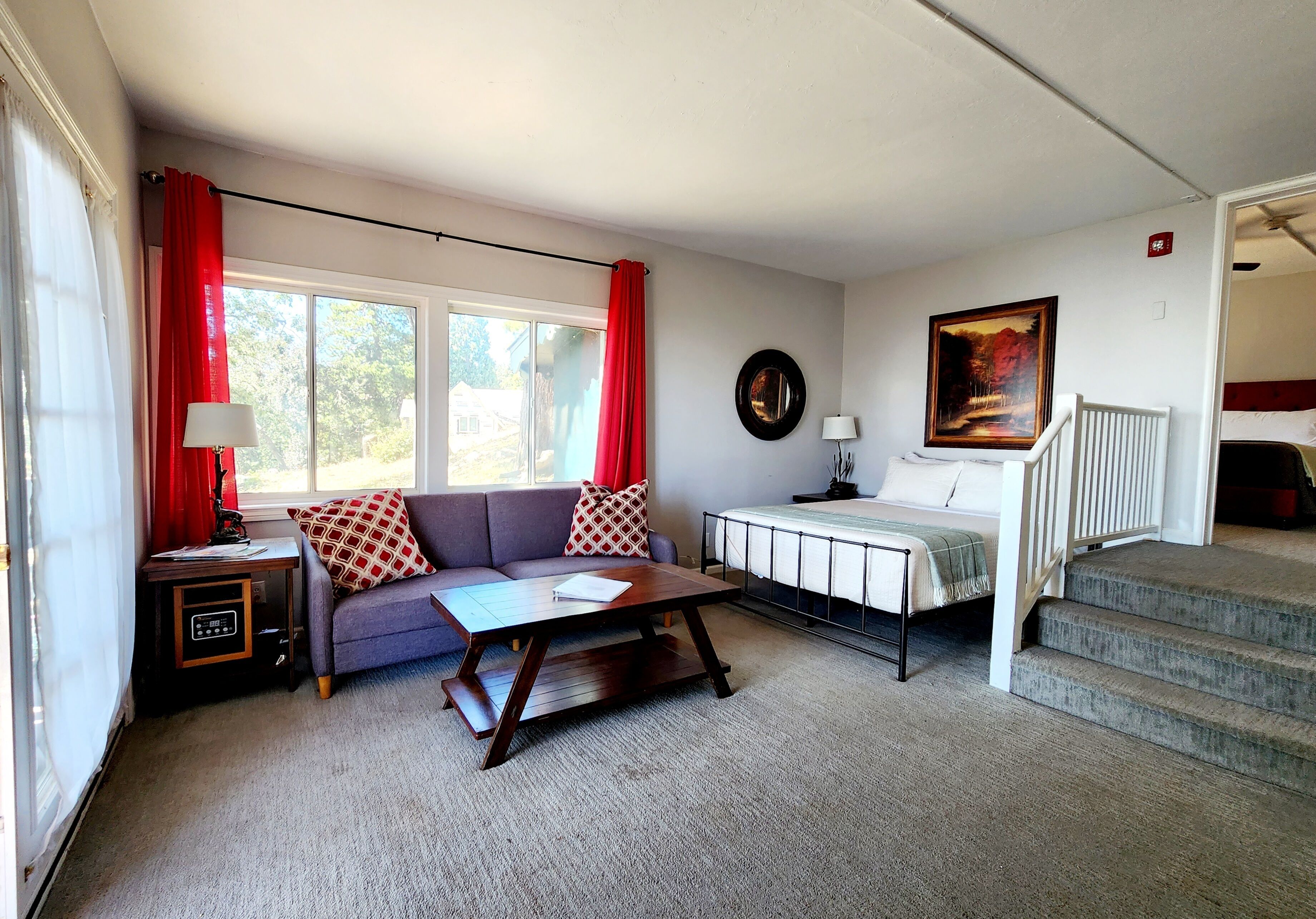 Una sala de estar con sofá, mesa de centro y cama con cabecera. Hay una ventana con cortinas, un cuadro en la pared y una escalera.