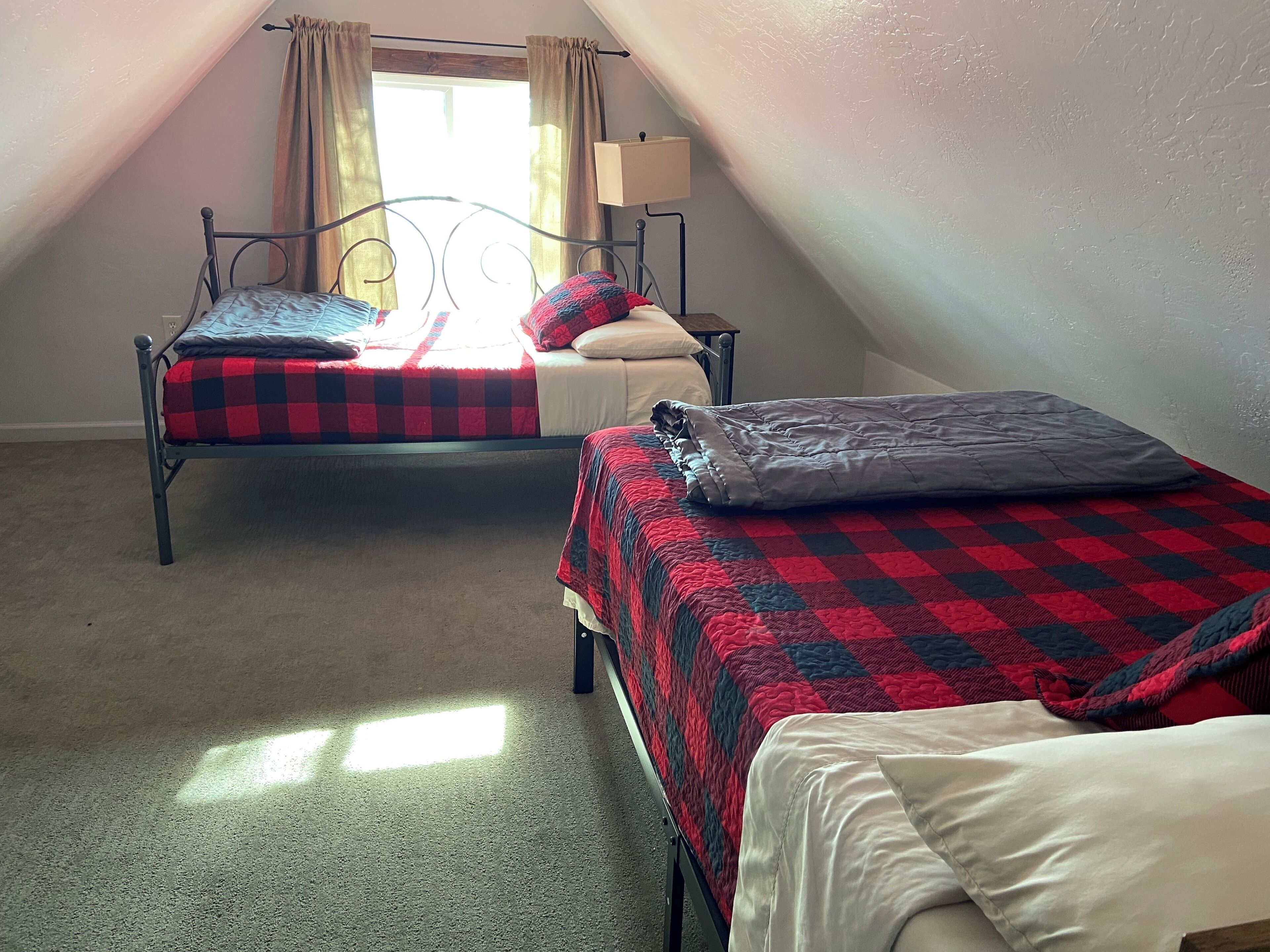 A room with two beds, each with a checkered blanket, a bedside table with a lamp, and a window with curtains.