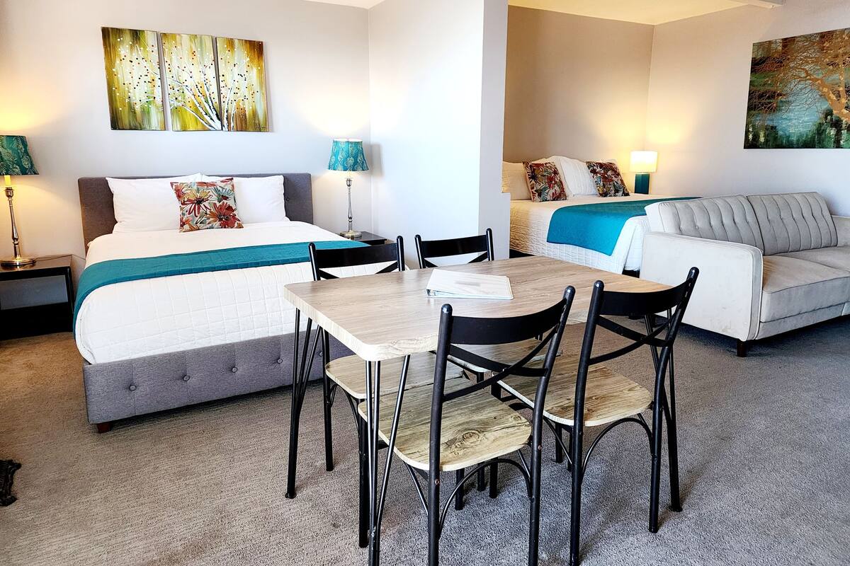 A hotel room with a bed, a sofa, a dining table with chairs, and artwork on the walls.