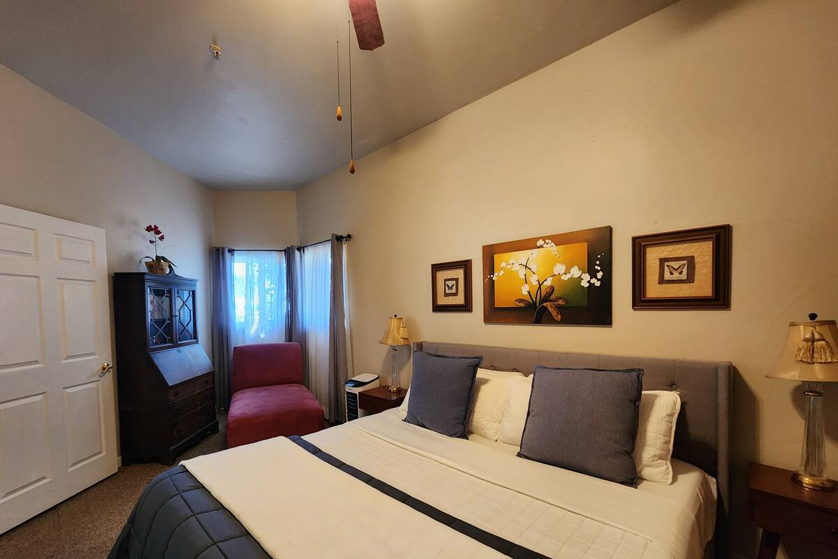 A bedroom with a bed, a nightstand, a lamp, a chair, a wardrobe, and a window with curtains.