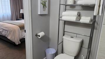 A bathroom with a toilet, a shelf with towels, a trash bin, and a toilet paper holder.
