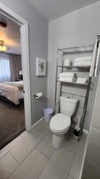 A bathroom with a toilet, a shelf with towels, a trash bin, and a toilet paper holder.