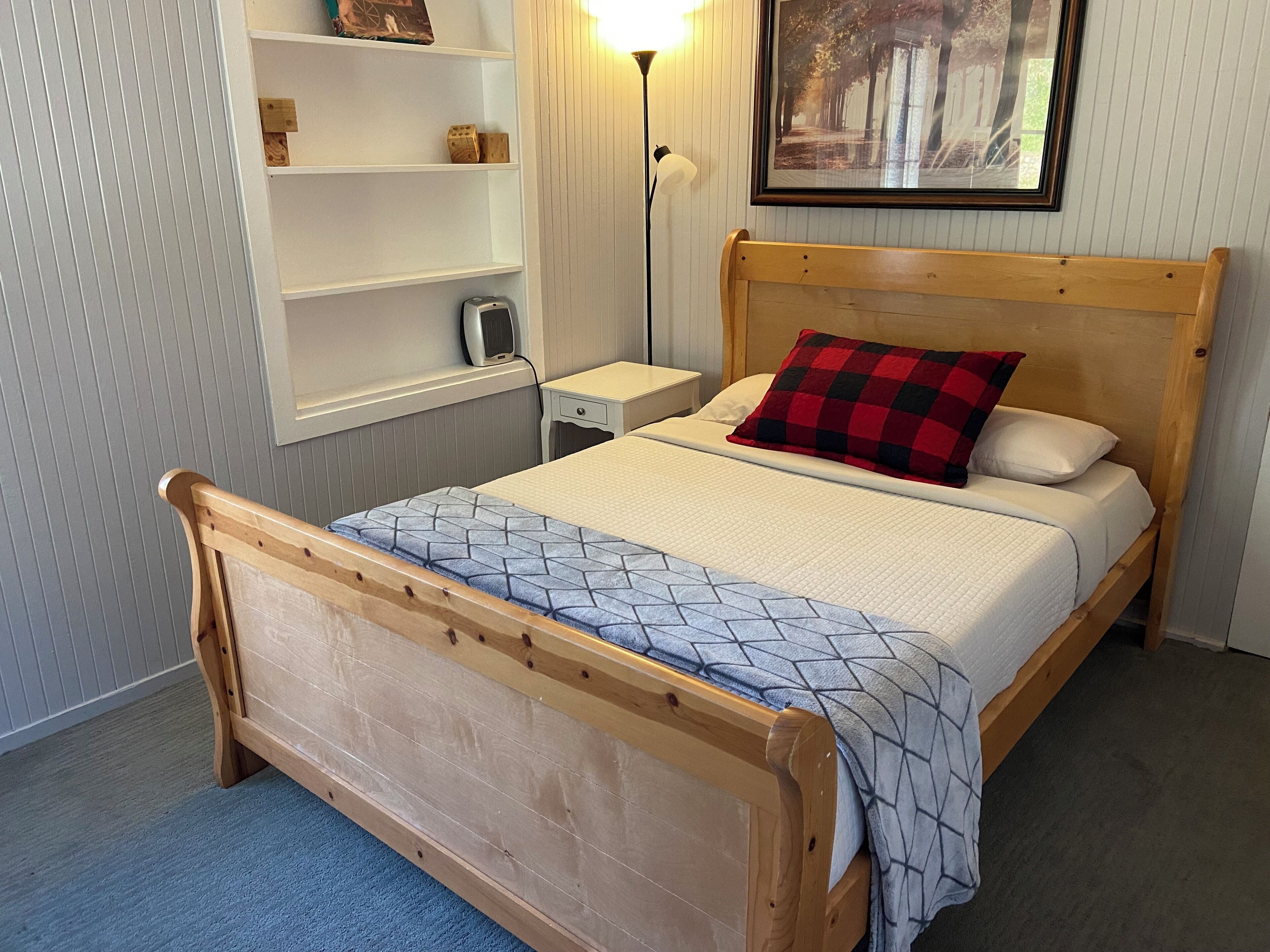 A wooden bed with a geometric-patterned bedspread and a red and black checkered pillow. There is a nightstand with a lamp, a shelf with decorative items, and a framed picture on the wall.