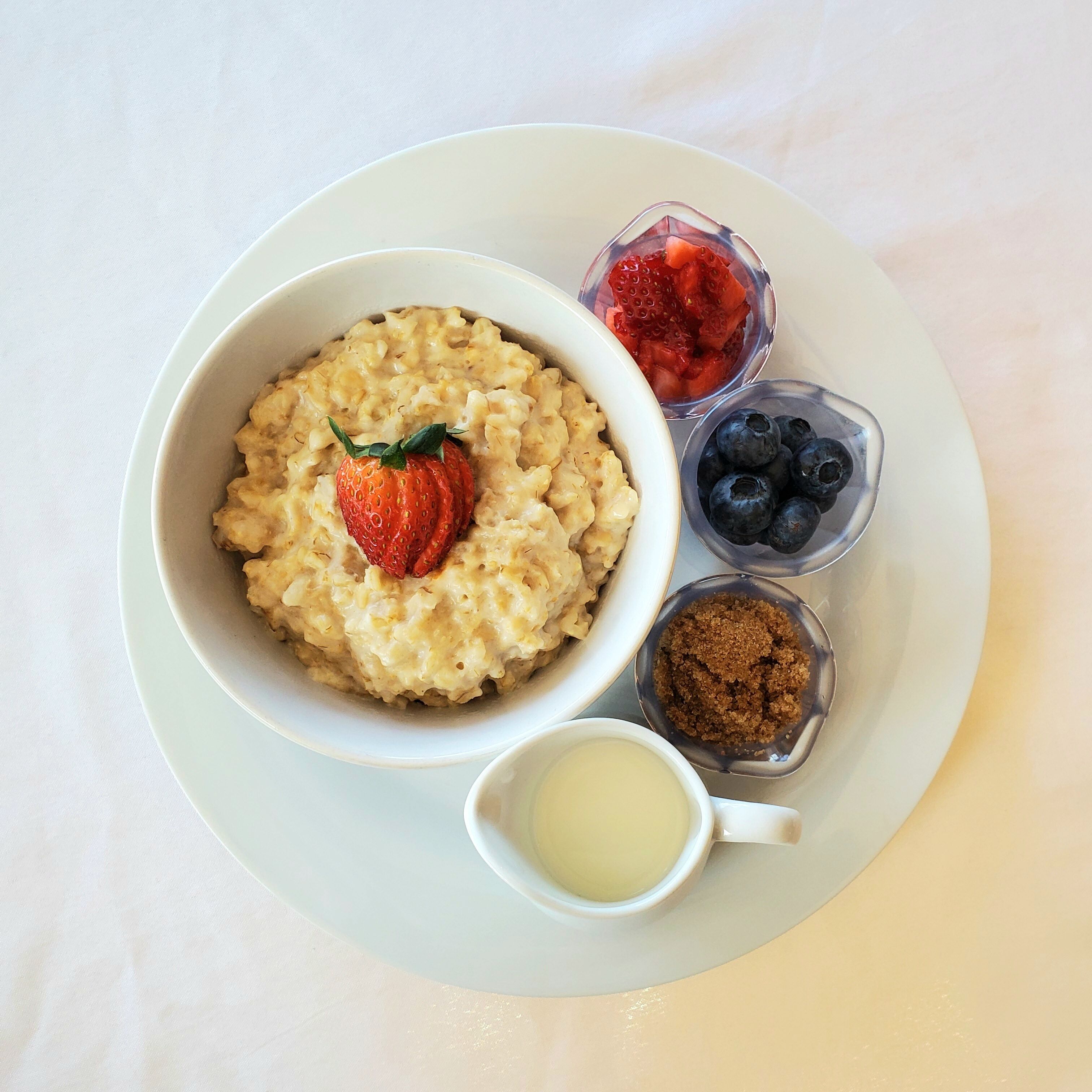 A bowl of oatmeal with a strawberry on top, accompanied by a small bowl of blueberries, a small bowl of brown sugar, and a small cup of milk.