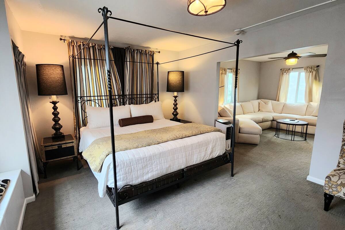 A four-poster bed with a canopy, a sofa, a chair, and a small table in a well-lit room with a carpeted floor.