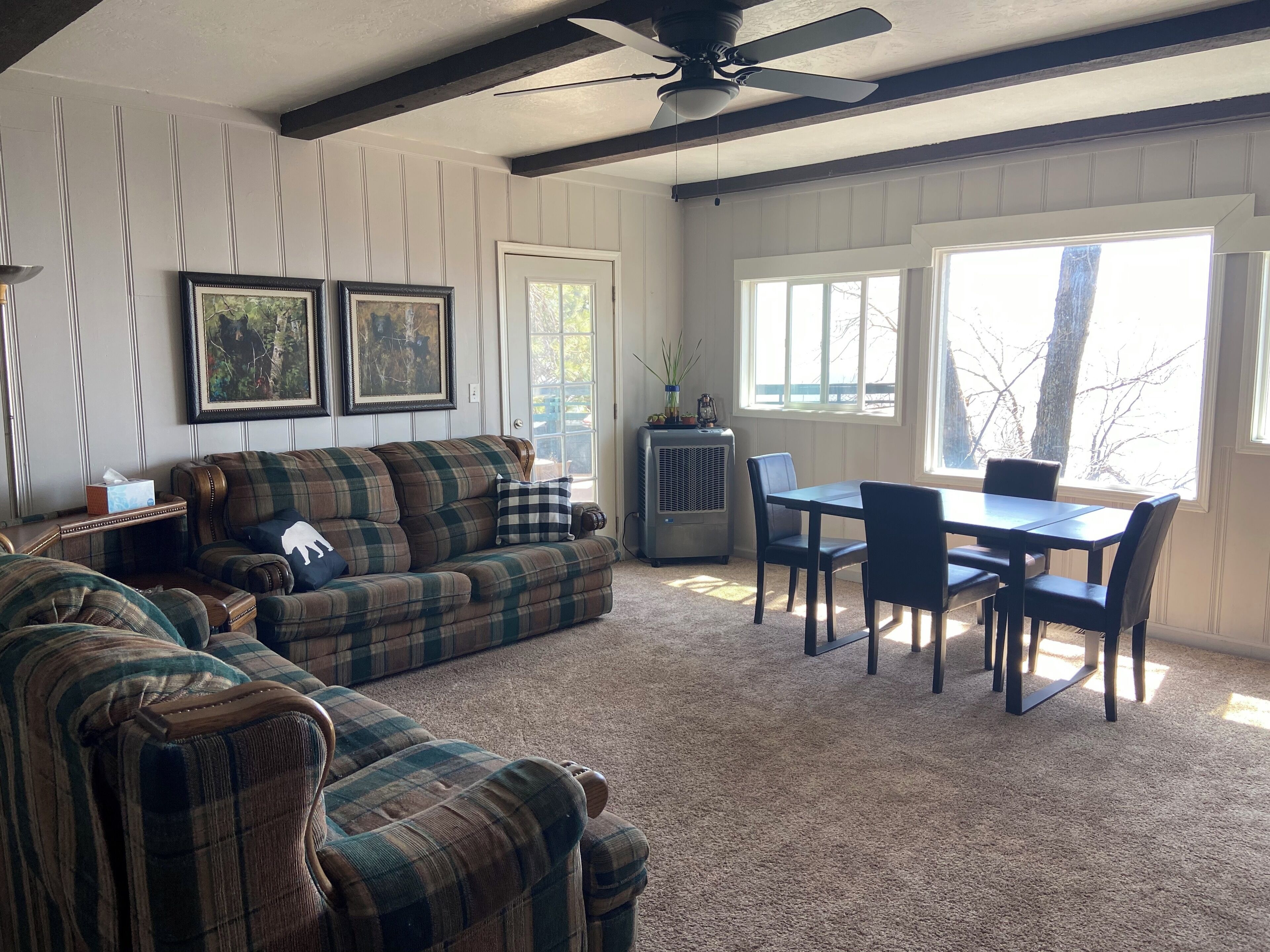 A living room with a plaid sofa, a dining table with chairs, a ceiling fan, and framed pictures on the wall.