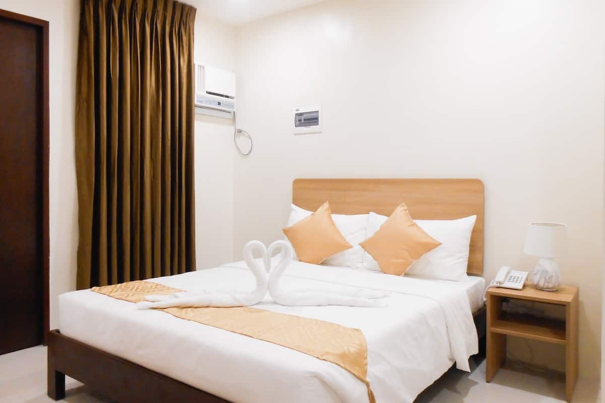 A hotel room with a wooden bed, white bedding, a brown throw, a nightstand with a lamp, and a wall-mounted air conditioner.