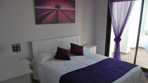 A bedroom with a bed, white headboard, two pillows, a purple blanket, a nightstand, a lamp, and a painting on the wall.
