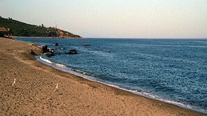A sandy beach with gentle waves, a clear sky, and a distant hill with a structure.