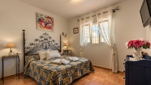 A bedroom with a patterned bedspread, a decorative headboard, a bedside table with a lamp, a dresser, and a window with curtains.