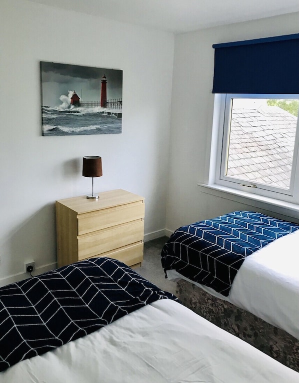 A bedroom with two beds, a dresser, a painting of a lighthouse, and a window with a view of the roof. - 3 bed house, free WiFi, off street parking. Central Ayrshire, 20 mins to Glasgow (KILMARNOCK, SCOTLAND)