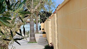 A narrow concrete pathway flanked by yellow walls and trees with green foliage. - Bungalow in complex with shared pool (CONIL DE LA FRONTERA)
