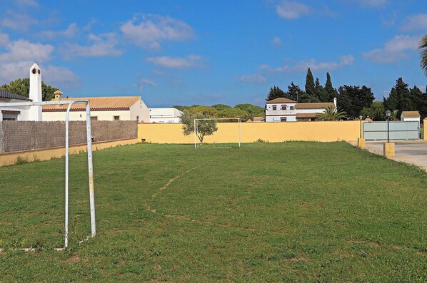 A grassy field with a goalpost, a white building with a red roof, and a yellow wall in the background. - Bungalow in complex with shared pool (CONIL DE LA FRONTERA)