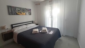 A neatly made bed with a dark quilt, two folded towels, a nightstand with a lamp, and a painting above the bed.