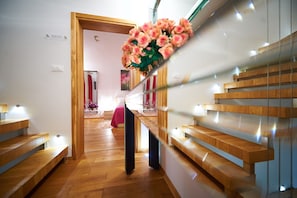 Un intérieur d’hôtel moderne avec un escalier en bois, un couloir central et un lit recouvert d’une couverture rose.