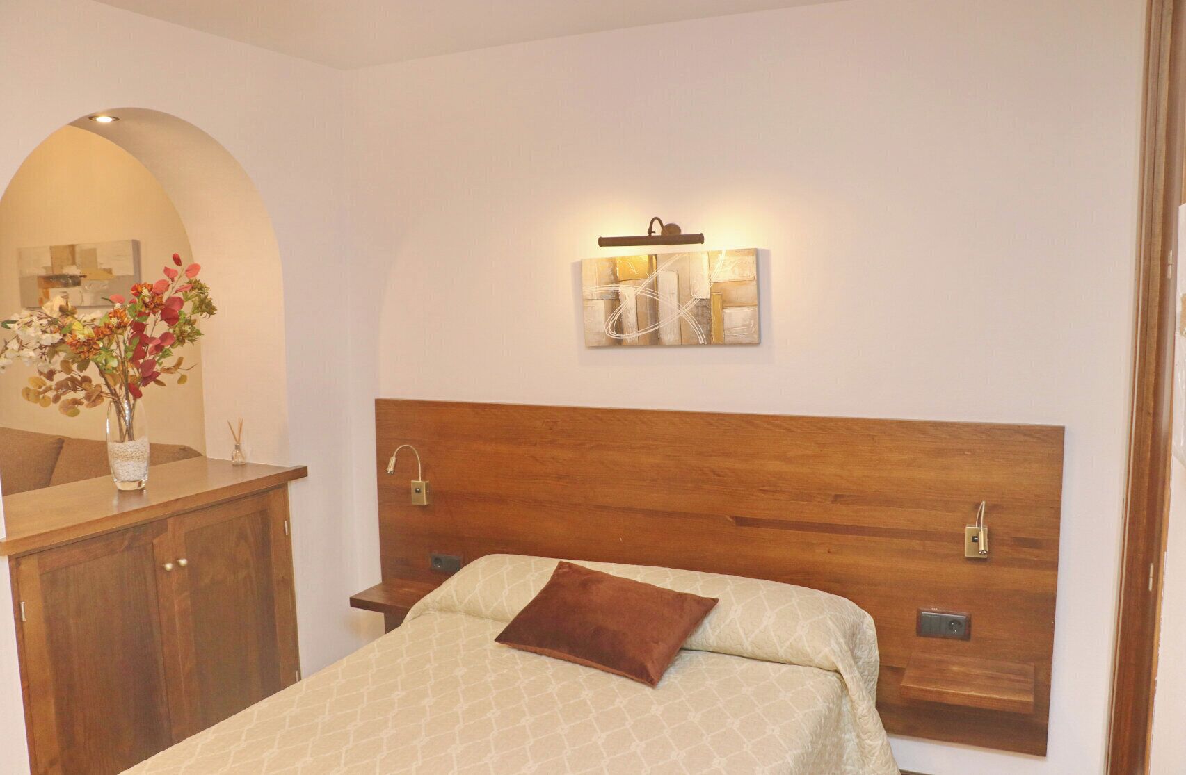 A bedroom with a wooden headboard, a bed with patterned bedding, a nightstand, a bedside table, a wall-mounted light, and a decorative piece above the bed.