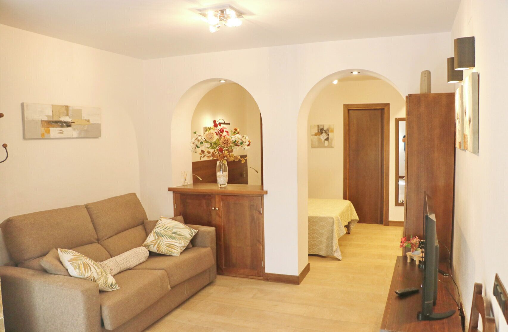 A living room with a sofa, a wooden cabinet, a TV, and a hallway leading to a bedroom.