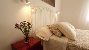 Ein Schlafzimmer mit einem weißen Metall-Kopfteil, einem Nachttisch mit Blumen in einer Vase und einem Bett mit gemusterten Betttextilien.