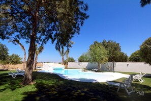 Une piscine avec des chaises longues et une pelouse, entourée d’arbres et d’un mur blanc.
