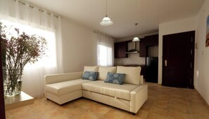 Ein Wohnzimmer mit einem beigefarbenen Ecksofa, zwei gemusterten Kissen, einer Vase mit Trockenblumen sowie einer Küche im Hintergrund und einer dunklen Holztür.