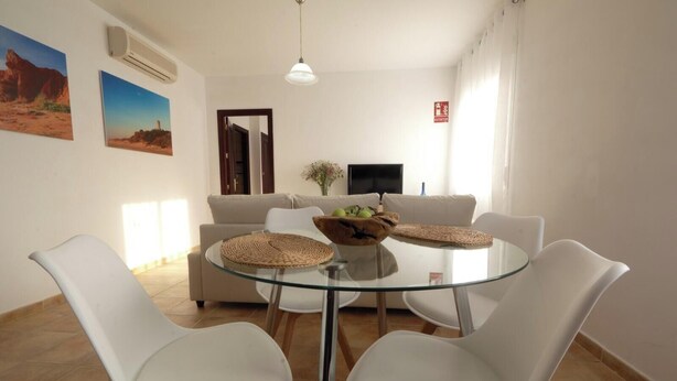 A dining area with a glass table, white chairs, a beige sofa, and a TV in the background.
