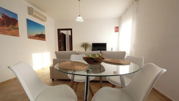 A dining area with a glass table, white chairs, a beige sofa, and a TV in the background.