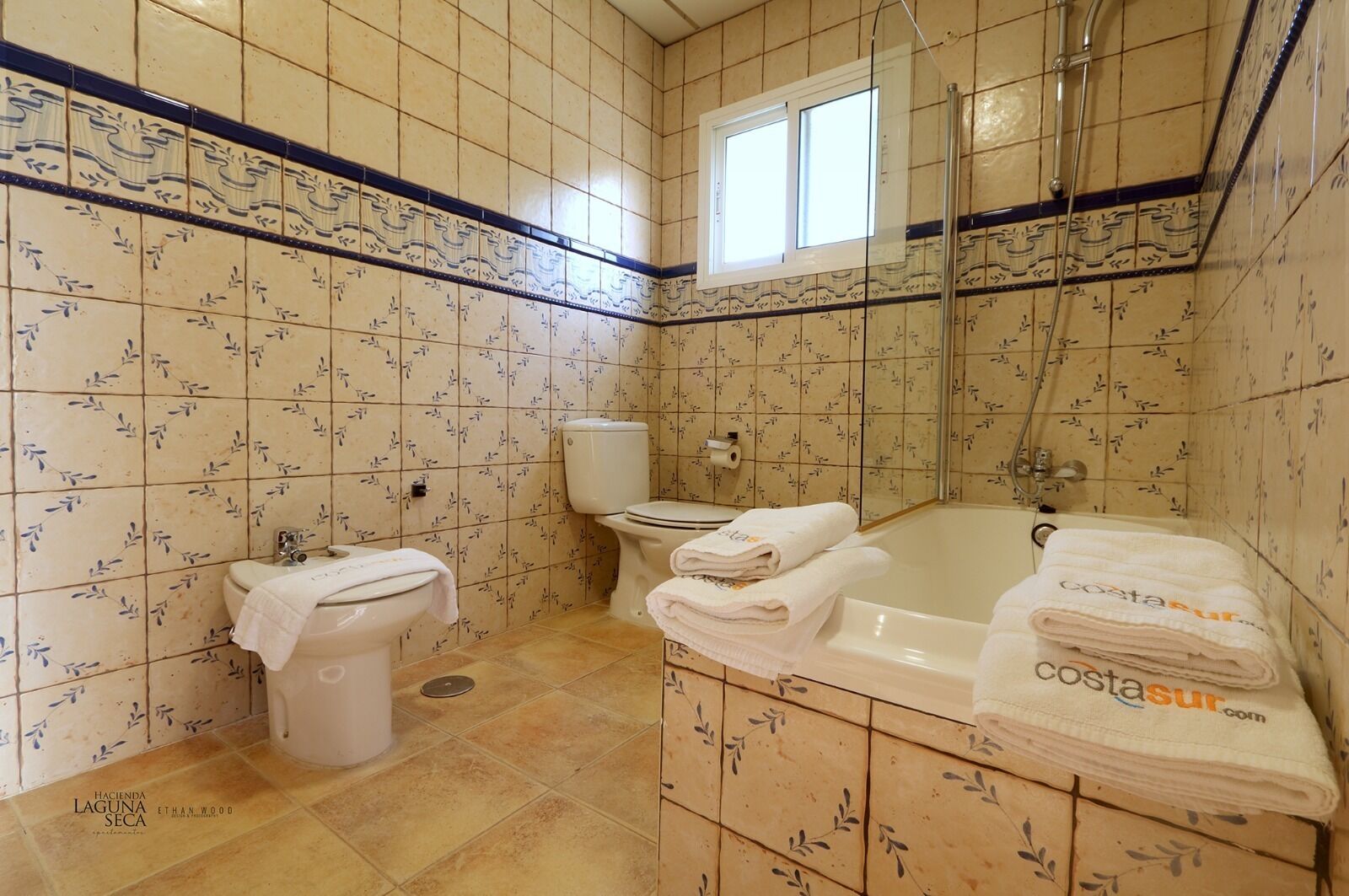 Une salle de bain avec des carreaux décorés, des toilettes, un lavabo, une baignoire avec des serviettes et une douche.