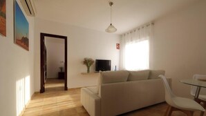 A living room with a sofa, a TV, a table with chairs, a window with curtains, and a door leading to another room.
