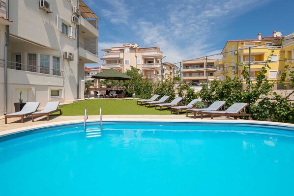 A swimming pool with lounge chairs, a grassy area, and buildings in the background.