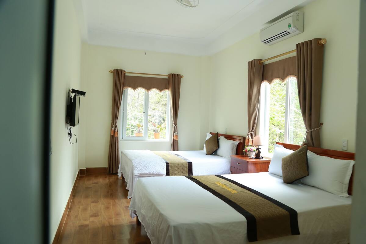 Quarto de hotel com duas camas, piso de madeira, janela com cortinas, aparelho de ar condicionado e mesa de cabeceira com abajur.