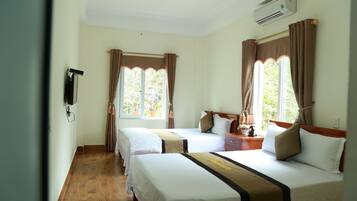 A hotel room with two beds, a wooden floor, a window with curtains, a bedside table with a lamp, and an air conditioning unit.