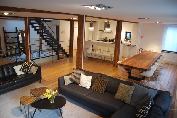 A spacious living area with a black sofa, a wooden dining table, and a staircase leading to an upper level.