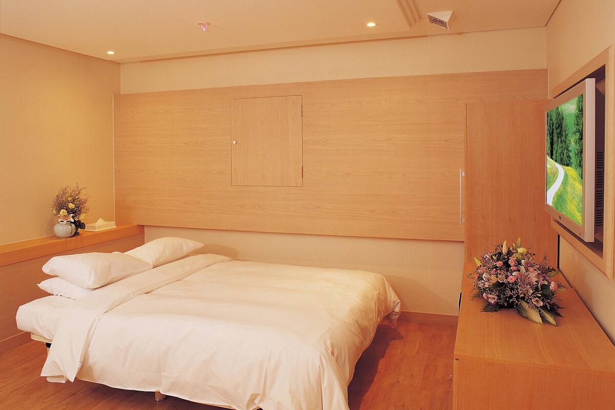A bedroom with a large bed, wooden headboard, a flat-screen TV mounted on the wall, and a small desk with a vase of flowers.