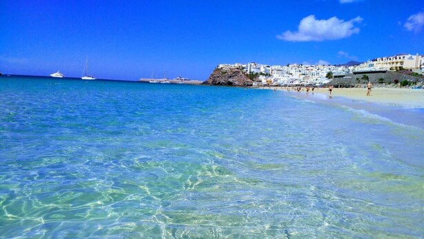 A coastal beach scene with clear turquoise water, a sandy shore, and a backdrop of white buildings. - Great location, apartment Fuerteventura (Costa Calma (Fuerteventura))