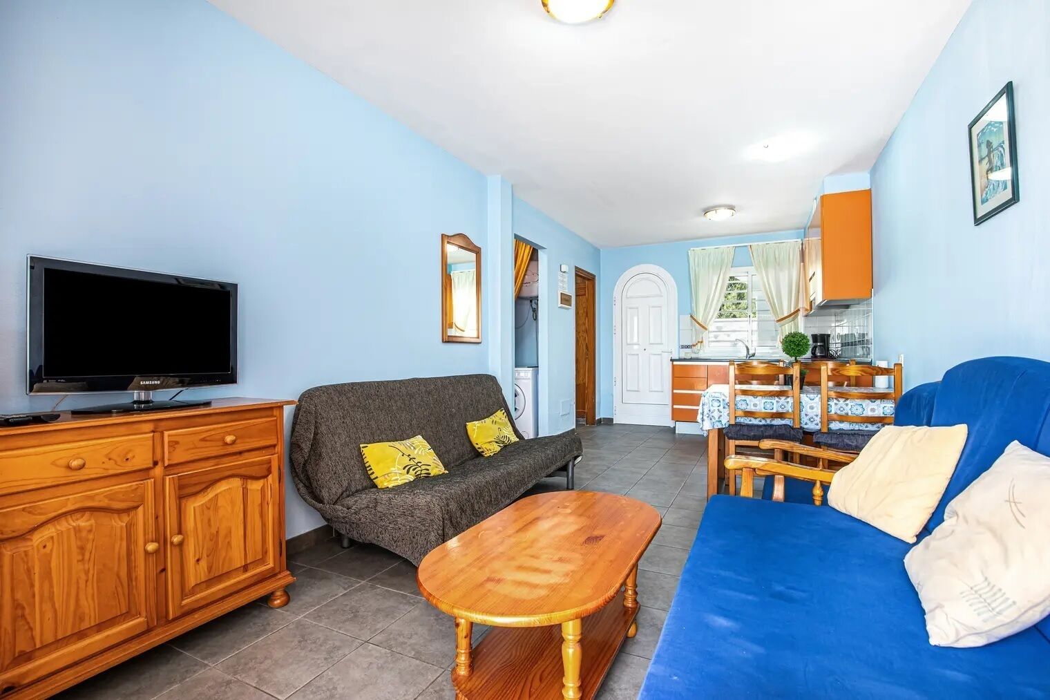 A living room with a TV, sofa, and wooden furniture. There is a kitchen in the background with a sink and cabinets.