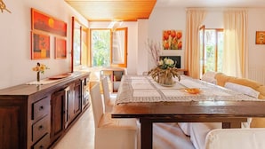 A dining area with a wooden table, white chairs, and a vase of flowers. There are paintings on the walls and a window with curtains.