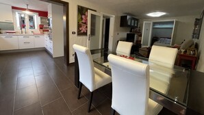 A modern kitchen with white cabinets and a red backsplash, a dining area with a glass table and white chairs, and a living room with a sofa and a TV.