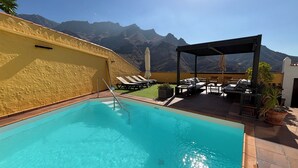A pool with clear blue water, surrounded by lounge chairs and a covered seating area, with mountains in the background.
