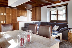 A cozy living area with a wooden dining table, leather chairs, a grey sofa, and a window with curtains.