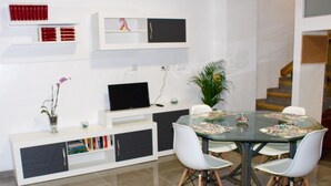 Una sala de estar moderna con una mesa de comedor de cristal, sillas blancas, un televisor de pantalla plana, una estantería y una escalera al fondo.