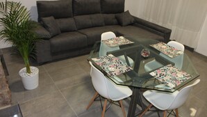 Una sala de estar moderna con una mesa de comedor de cristal rodeada por sillas blancas, un sofá gris oscuro y una planta en maceta.