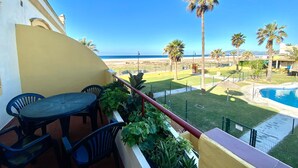 Un balcón con mesa y sillas, una piscina y vistas a la playa y palmeras.