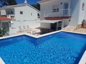 Una piscina con borde de azulejos, un patio techado con sillas y mesas, y un edificio blanco con balcón.