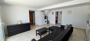 Una sala de estar moderna con un sofá negro, una mesita, un mueble negro y una zona de cocina con refrigerador y lavadora.