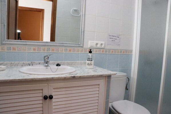 Un baño con lavamanos, espejo e inodoro. Hay un cartel en la pared y un dispensador de jabón en el lavamanos.