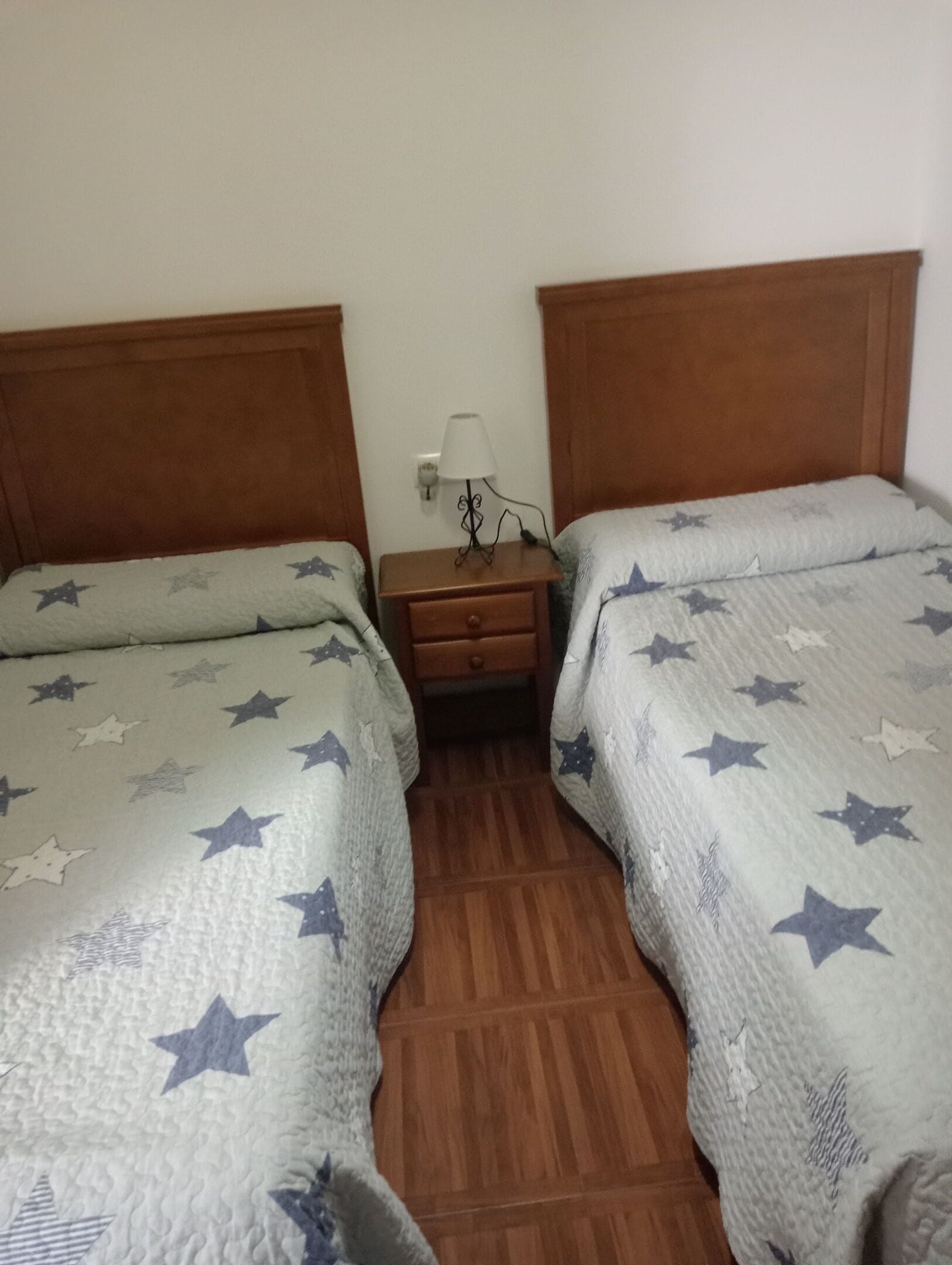 A room with two beds, each with a star-patterned bedspread, a wooden headboard, and a nightstand with a lamp between the beds.