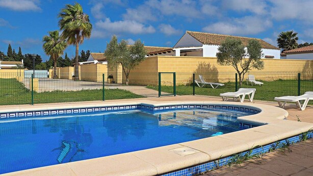A swimming pool with a tiled edge, surrounded by a fence, and adjacent to a building with a tiled roof.