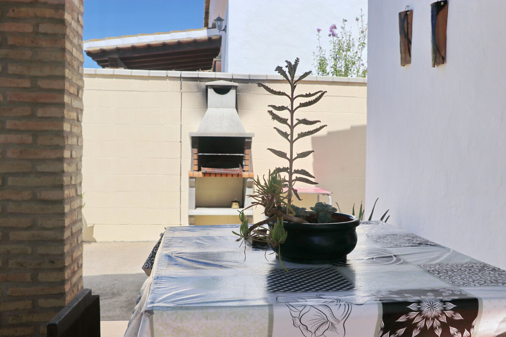 A patio with a table covered in a patterned tablecloth, a potted plant, and a brick wall.